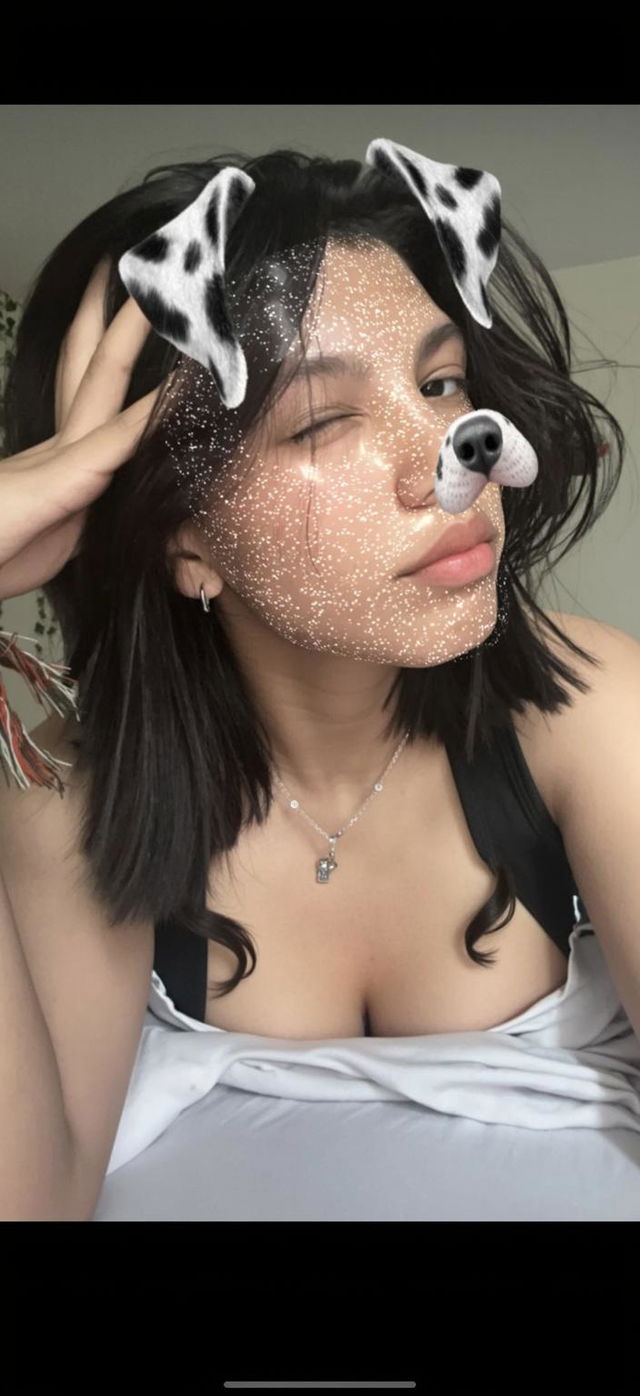 A young woman with short black hair, wearing a black sleeveless top, has a digital dog filter overlay on her face featuring dog ears and a nose