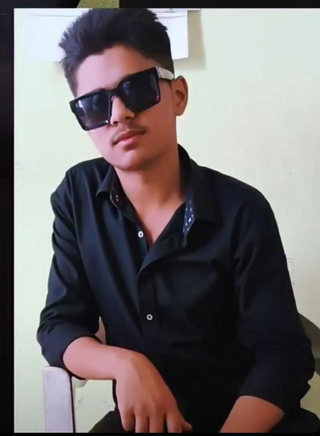 A young man with a relaxed pose, wearing large sunglasses and a black shirt, sitting on a chair