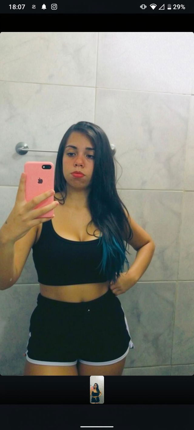 A woman taking a selfie in a bathroom mirror, wearing a black tank top and black shorts with white trim