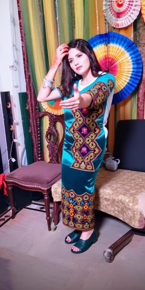 A woman in a deep blue traditional outfit with intricate silver embroidery, standing in a festive room decorated with colorful paper fans and curtains