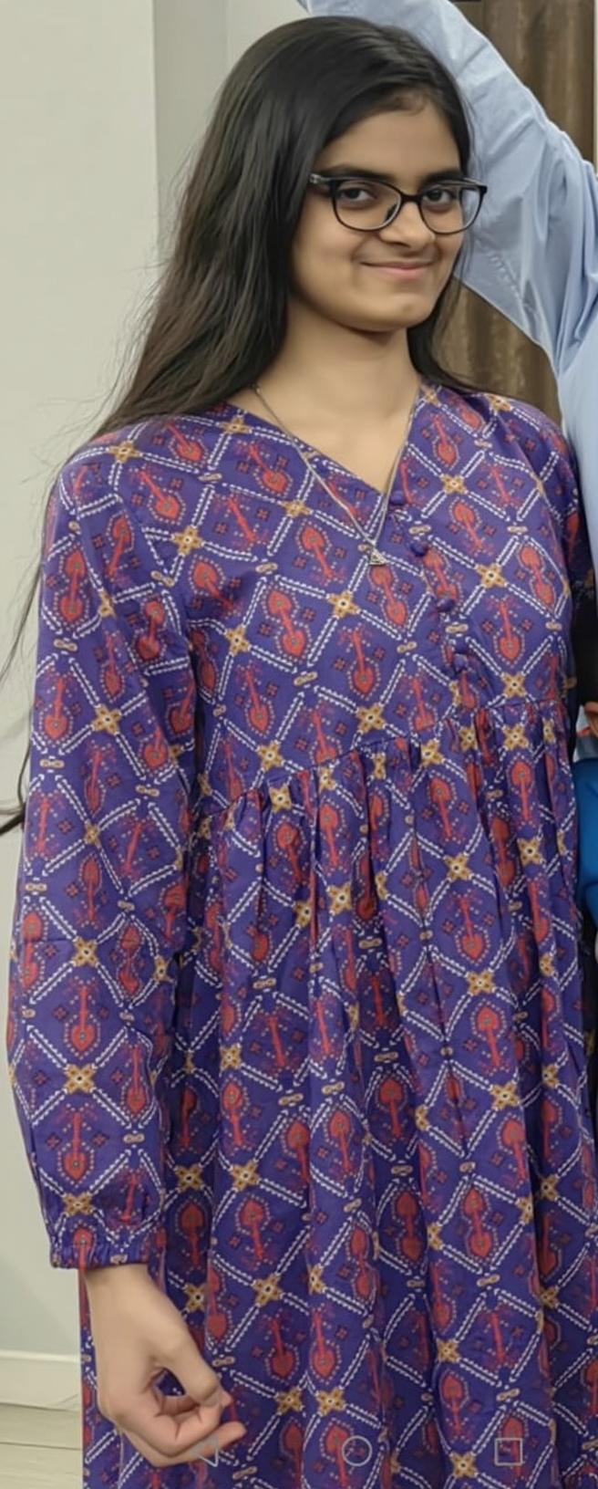 A person standing indoors wearing a long, vibrant purple dress with a unique geometric and paisley design