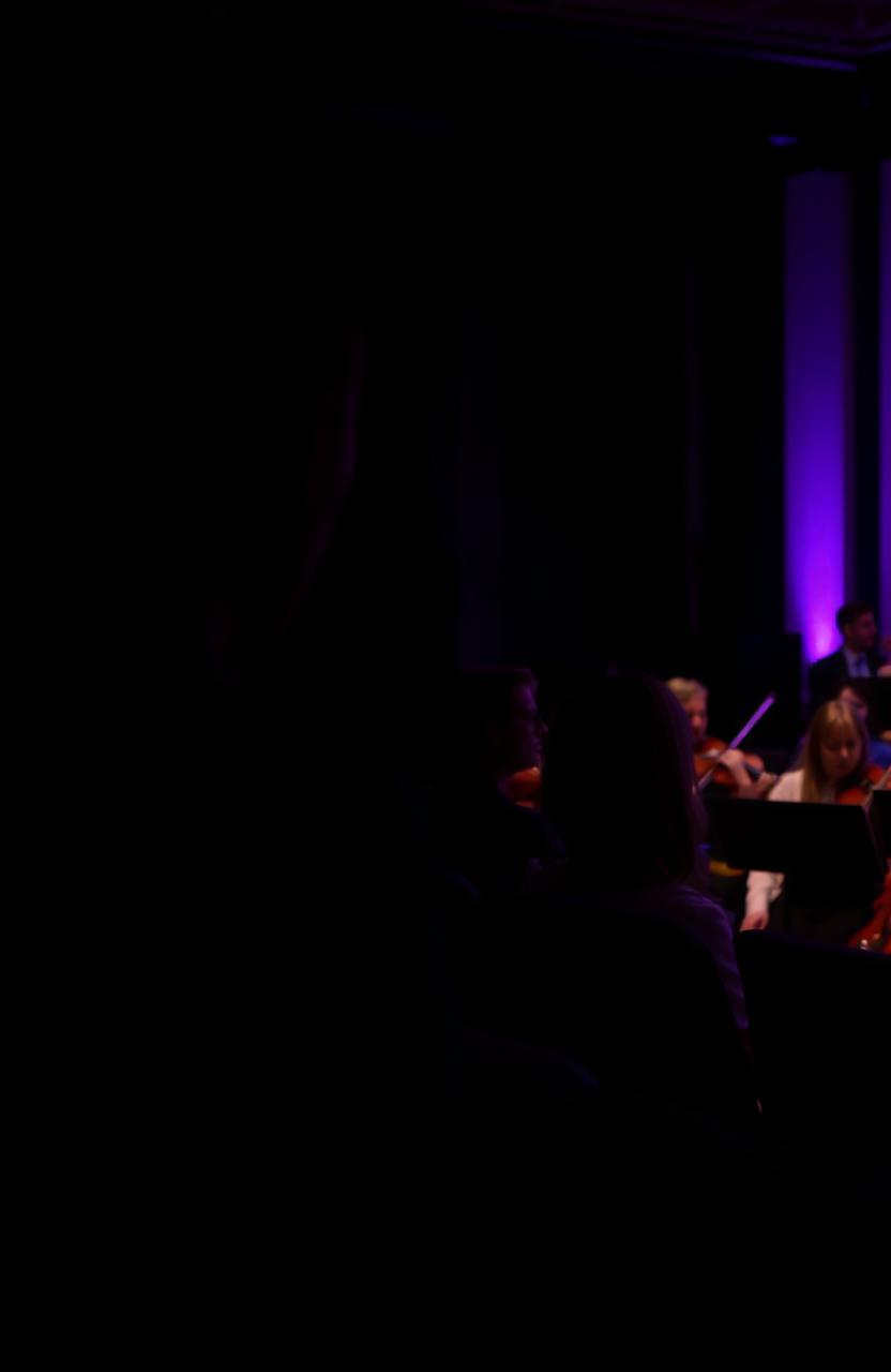 A silhouette of a man hiding in the distance, gazing towards the stands at a woman watching an orchestra