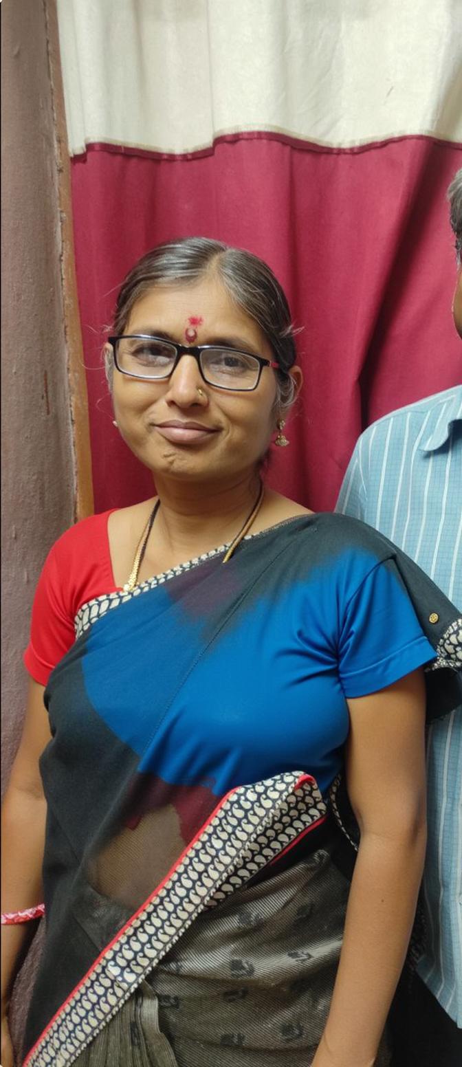 A woman wearing a blue blouse with a black and patterned sari, standing indoors with a neutral expression