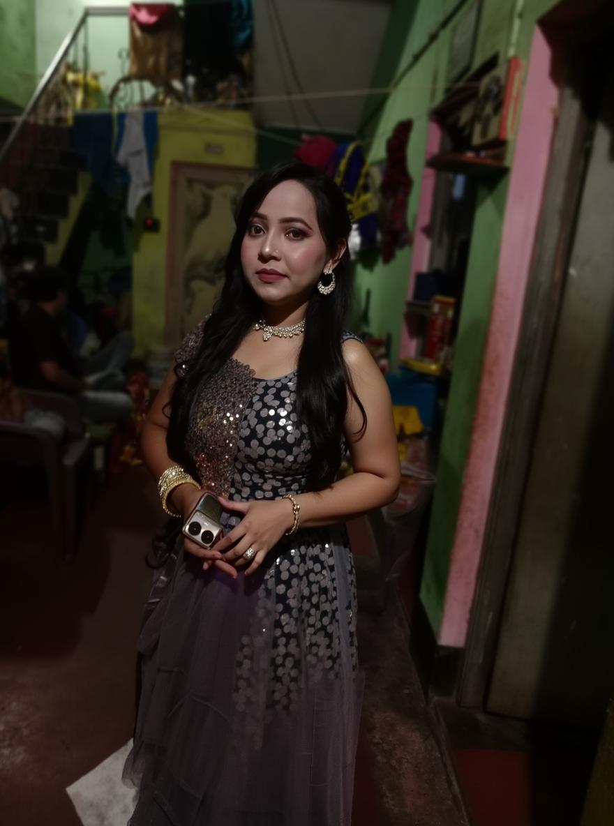 A glamorous woman posing in an elegant grey sequin gown, adorned with intricate beadwork and a flowing cape, standing in a colorful yet slightly rustic indoor setting