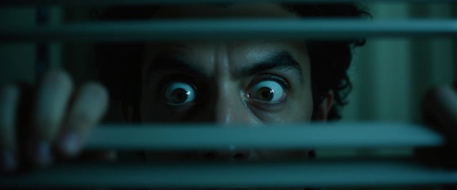 A captivating horror-themed image focused on a man's face as he lifts a blind, peering through it with wide, frightened eyes