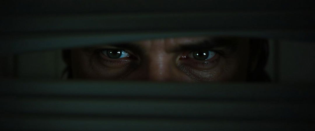 An intense close-up image of Jeremy Irons as he lifts a blind, peering through it with a look of deep fear in his eyes