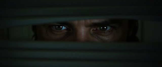 An intense close-up image of Jeremy Irons as he lifts a blind, peering through it with a look of deep fear in his eyes