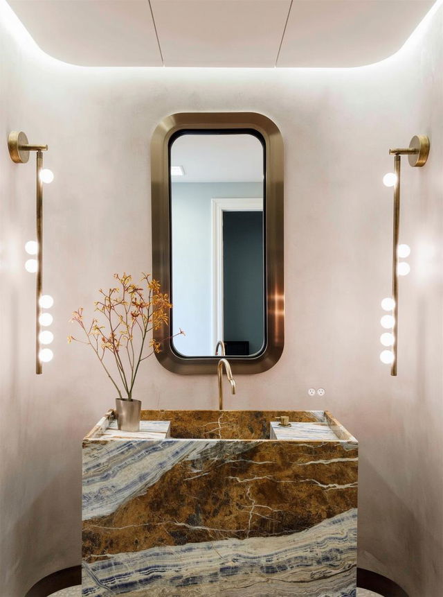 An elegant bathroom featuring a large, modern oval mirror with a sleek gold frame hanging above a stunning marble sink, characterized by rich brown and blue veins