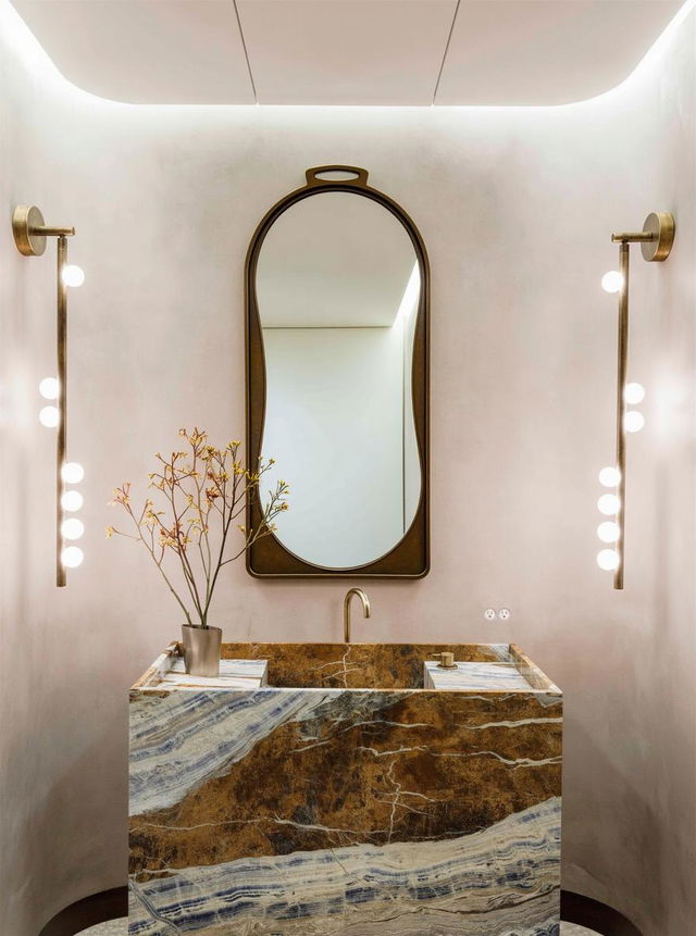 A chic bathroom interior featuring a large, modern oval mirror elegantly framed in gold, positioned above a stunning stone sink with rich brown and gold marbling