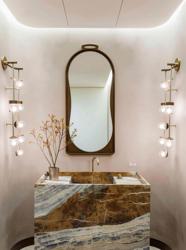 A sleek, modern bathroom showcasing a large, elongated oval mirror framed in gold, positioned above a striking marble sink featuring rich hints of brown and blue