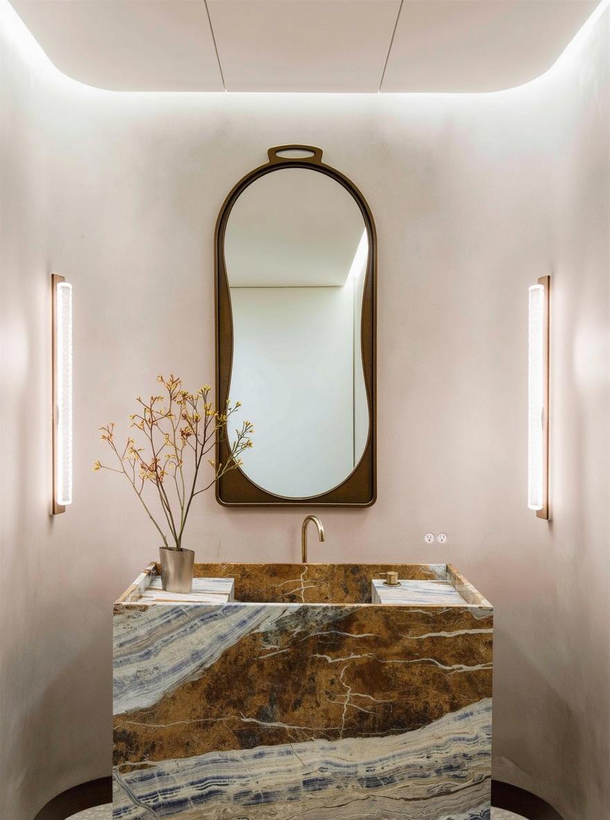 A stylish bathroom featuring a large, elegant mirror with a unique shape, framed in a sleek gold border, positioned above a stunning marble sink with deep brown and gold hues
