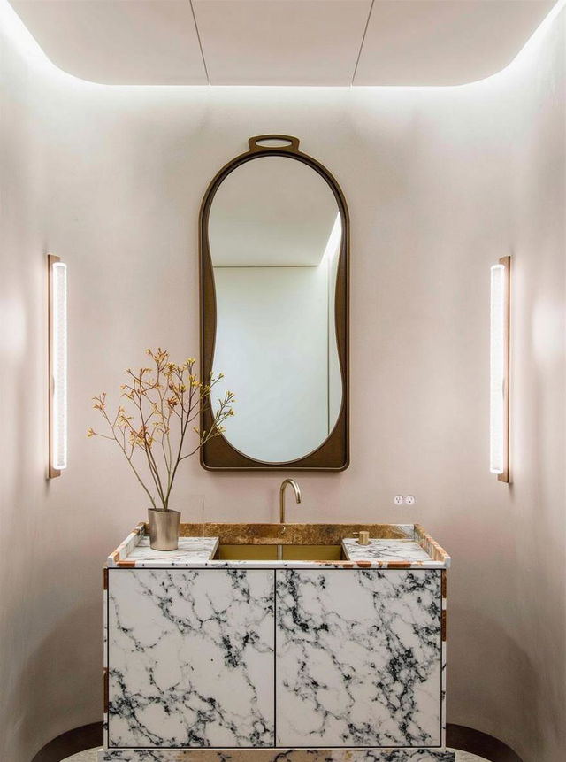 An elegant bathroom featuring a large oval mirror with a sleek gold frame, positioned above a beautifully textured sink made of smooth, creamy white marble with subtle gray veining