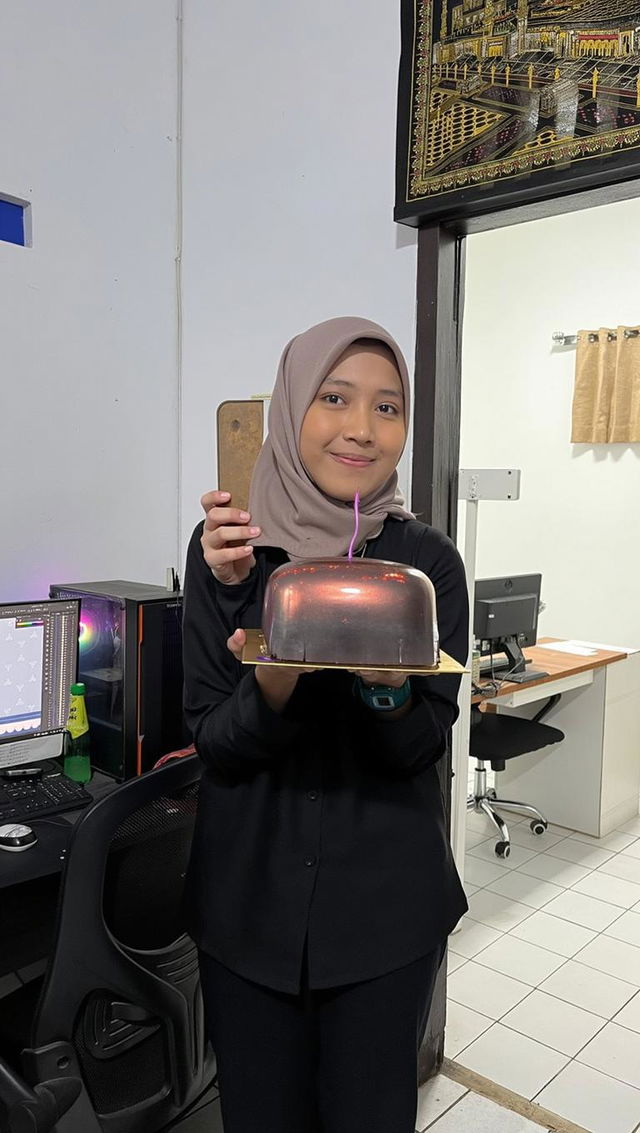 A young woman wearing a hijab, standing in an office environment, joyfully holding a beautifully decorated chocolate cake topped with whipped cream and cherries