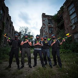 Five men dressed in tactical paintball gear, holding vibrant paintball guns, are gathered outside an eerie, abandoned sanatorium