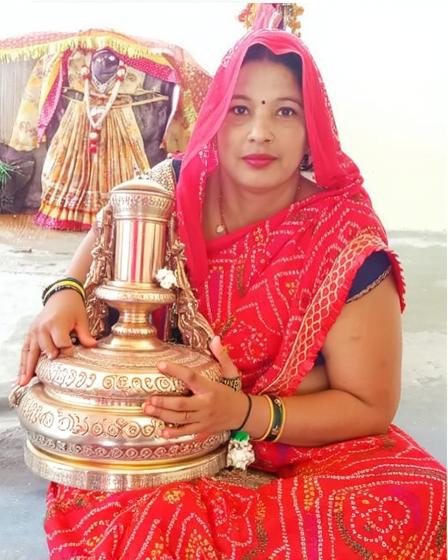 A woman wearing a vibrant red saree adorned with intricate golden patterns, holding a small decorative idol of a Hindu deity styled in bright, colorful clothing