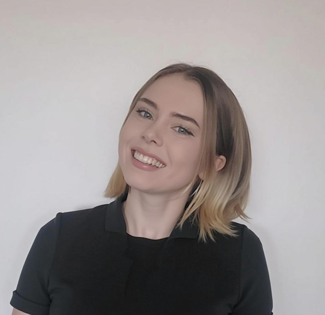 A confident young woman in a professional black blazer, smiling warmly with crossed arms