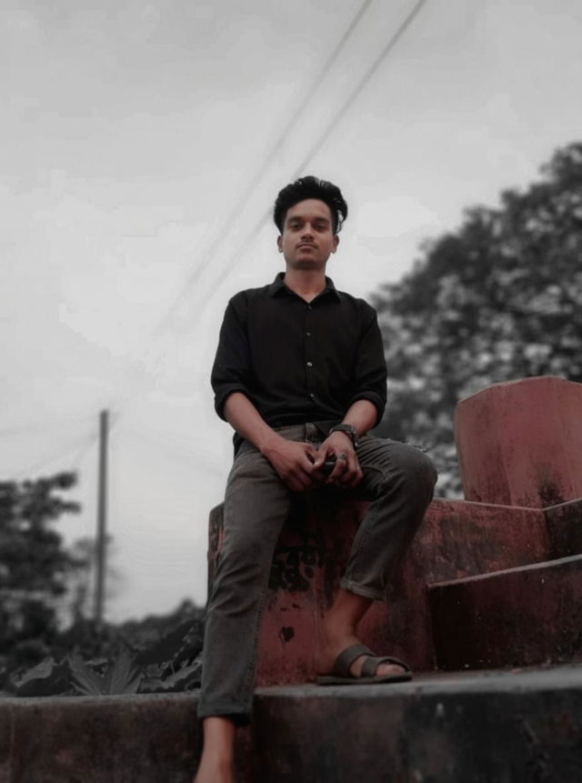 A stylish young man sitting casually on a red stone structure, wearing a black button-up shirt and faded jeans