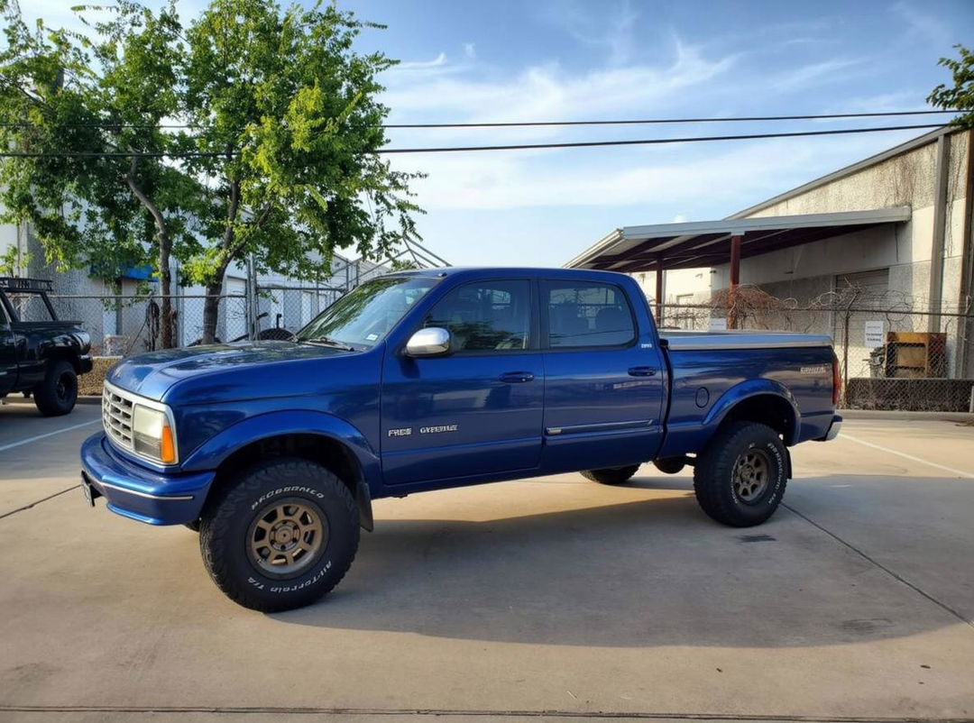 A blue pickup truck in a parking lot with the front of a 2000 Land Cruiser 100 seamlessly integrated into its design