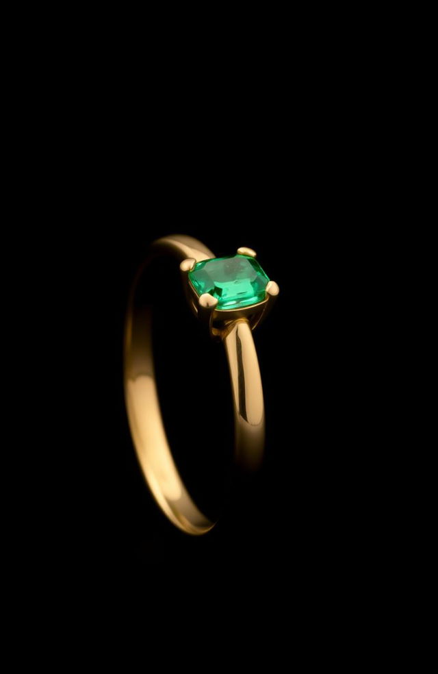 A simple golden ring with an emerald stone, positioned sideways, set against a mysterious black background