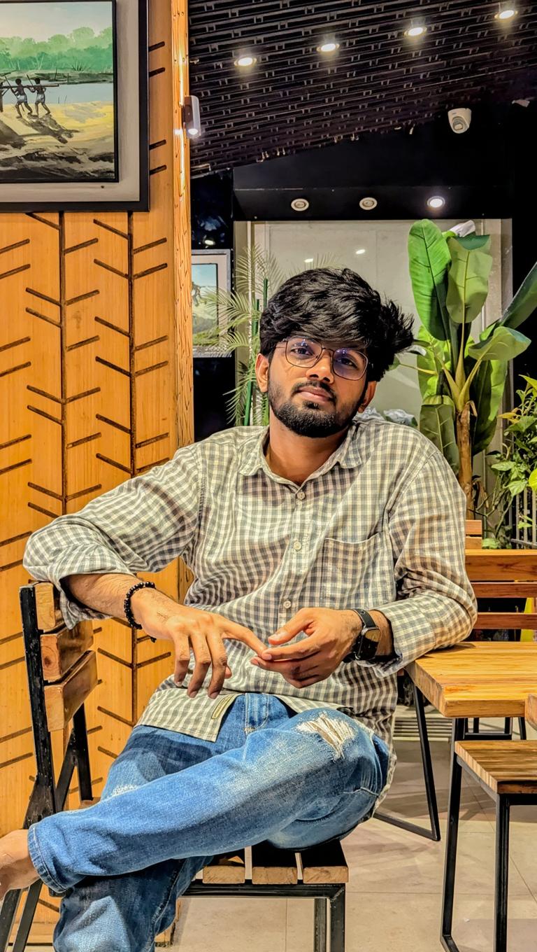 A young man sitting casually in a modern restaurant, wearing a checked shirt and distressed jeans