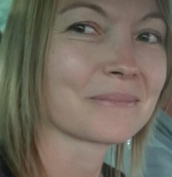 A close-up portrait of a smiling woman with medium-length blonde hair, sitting inside a vehicle with a soft, natural light illuminating her face