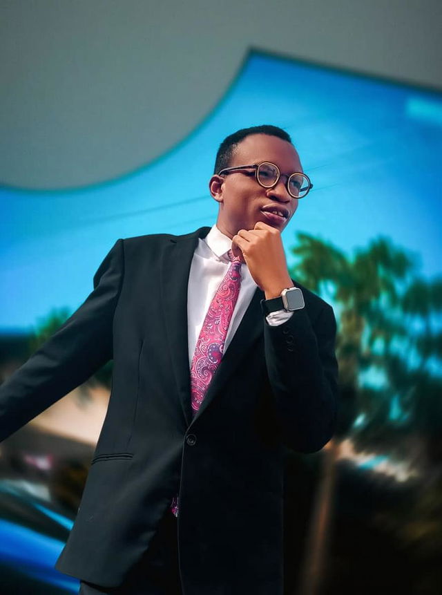 A young man confidently posing outdoors, wearing a classic black suit with a bright pink tie, glasses accentuating his stylish look