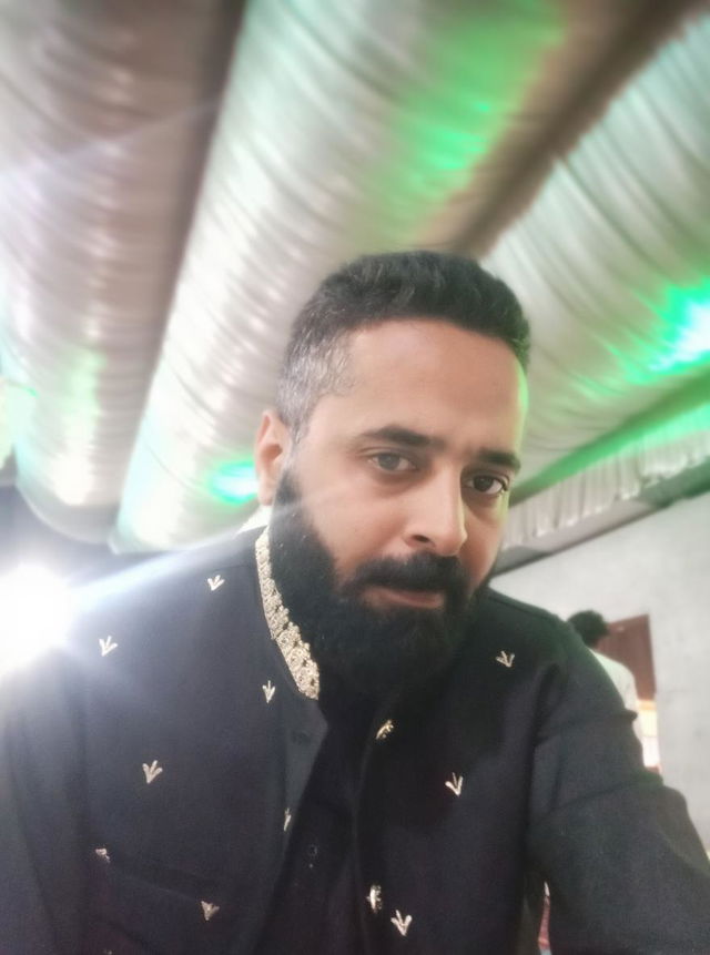 A well-dressed man with a beard, wearing a traditional black outfit with intricate golden embroidery, standing indoors with a softly lit ambiance