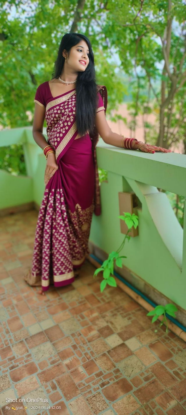 A beautiful woman wearing a traditional burgundy saree with golden embroidery, elegantly posed on a balcony surrounded by lush green foliage