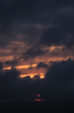 A stunning yet gloomy cloudy sunset sky filled with thick, dark gray clouds swirling together as the sun sets in the background, casting a subtle glow of muted orange and deep purple hues across the sky