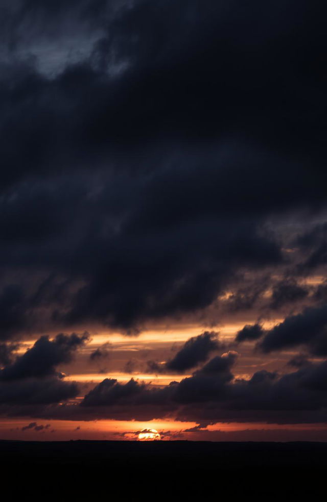 A stunning yet gloomy cloudy sunset sky filled with thick, dark gray clouds swirling together as the sun sets in the background, casting a subtle glow of muted orange and deep purple hues across the sky