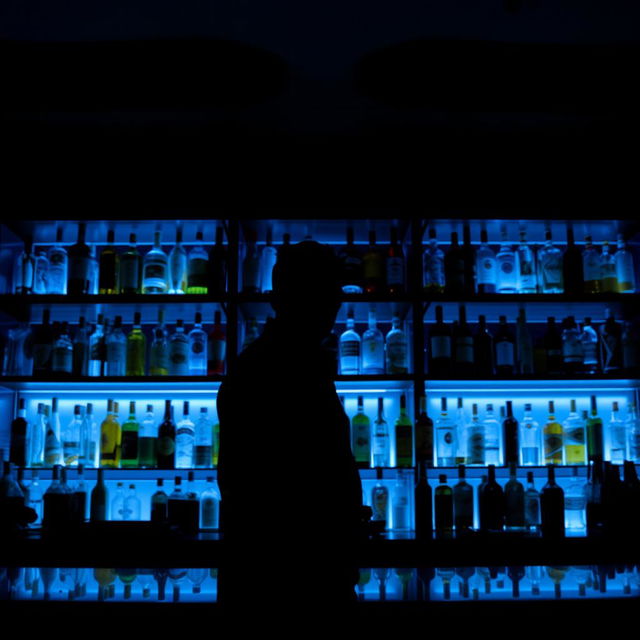 A bar scene showcasing a counter with shelves filled with a variety of bottles, illuminated by soft blue lights