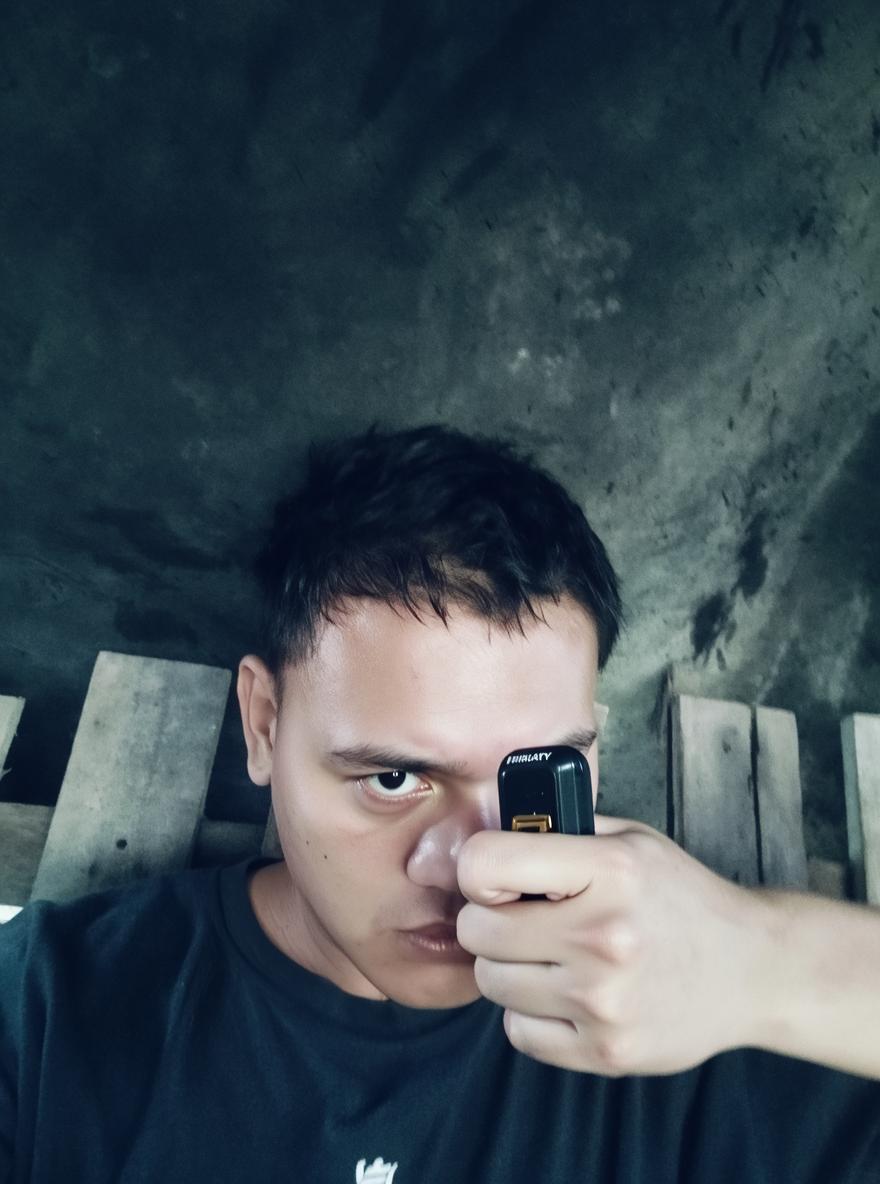 A close-up portrait of a young man with short, dark hair, looking intensely at the camera while holding a hand in front of his face, mimicking the shape of a gun