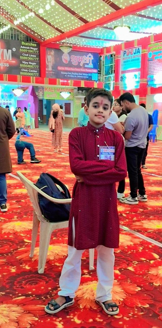 A cinematic scene set in a vibrant, festive hall with colorful decorations, featuring a young boy standing confidently in a traditional outfit, arms crossed