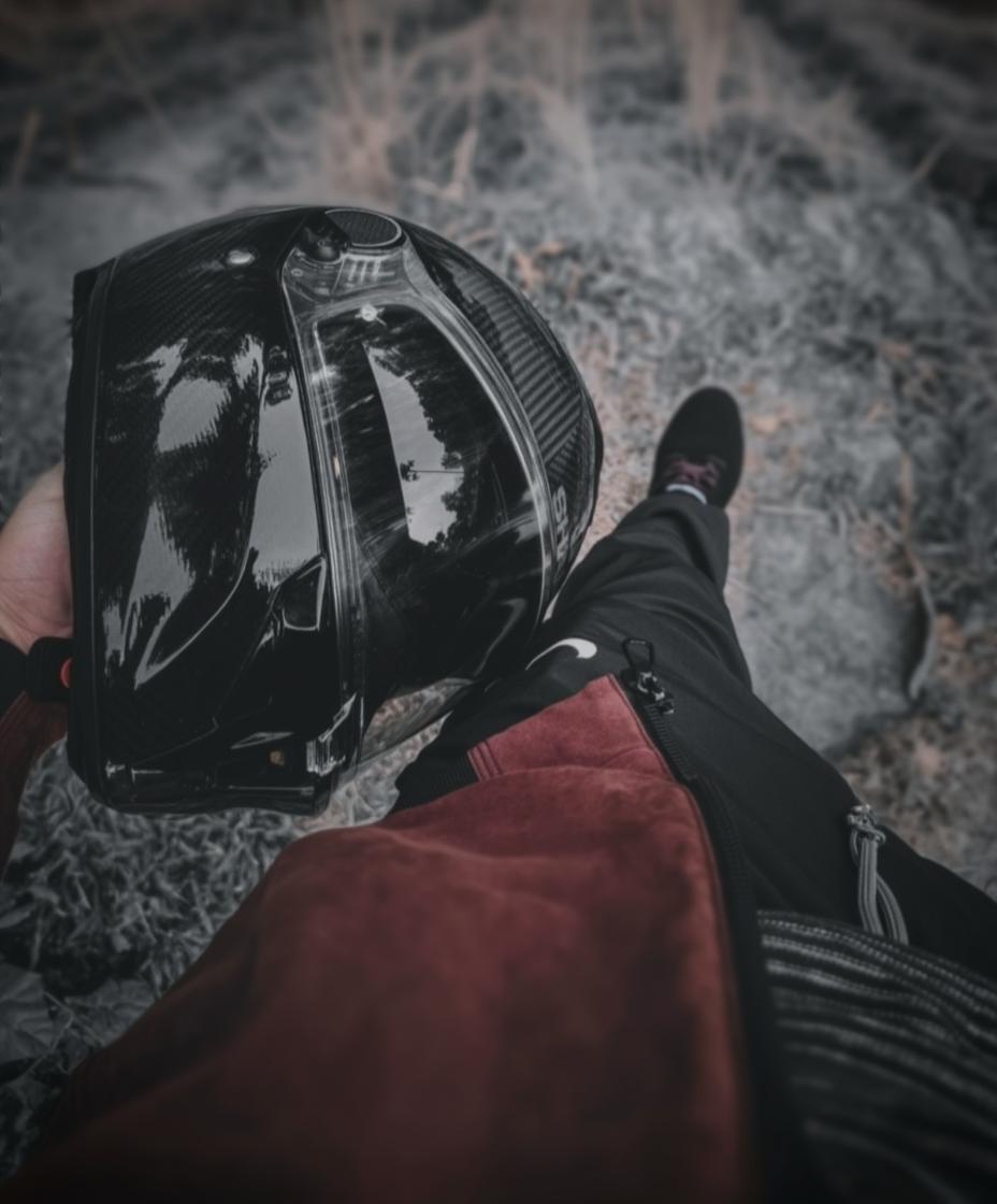 A close-up view of a person holding a sleek, black motorcycle helmet in one hand while standing on a rocky, natural landscape