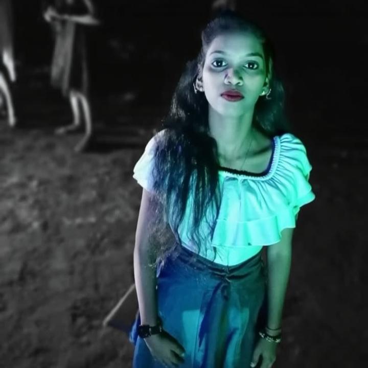 A young woman standing confidently in a dimly lit outdoor setting, wearing a charming off-the-shoulder white blouse and a flowing blue skirt