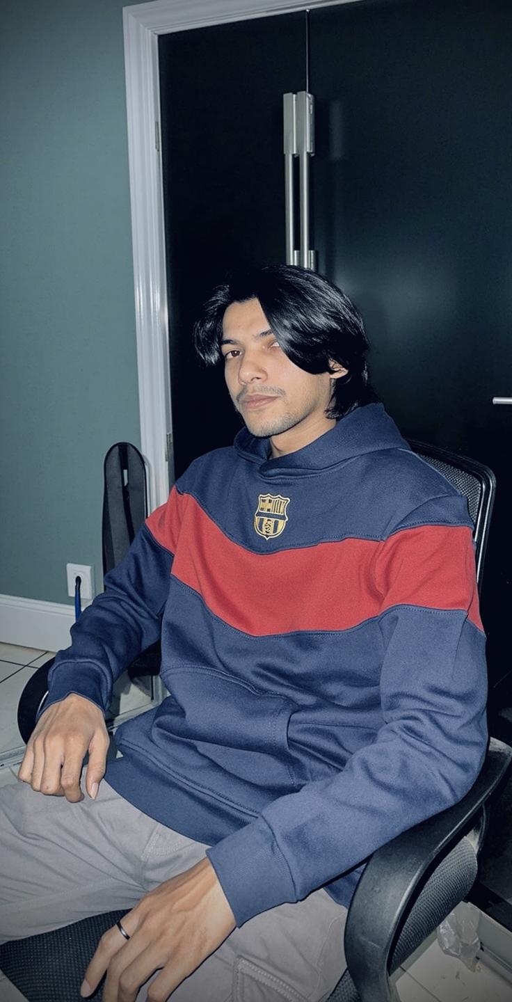 A young man with long black hair wearing a navy blue and red striped hoodie, seated in a black chair