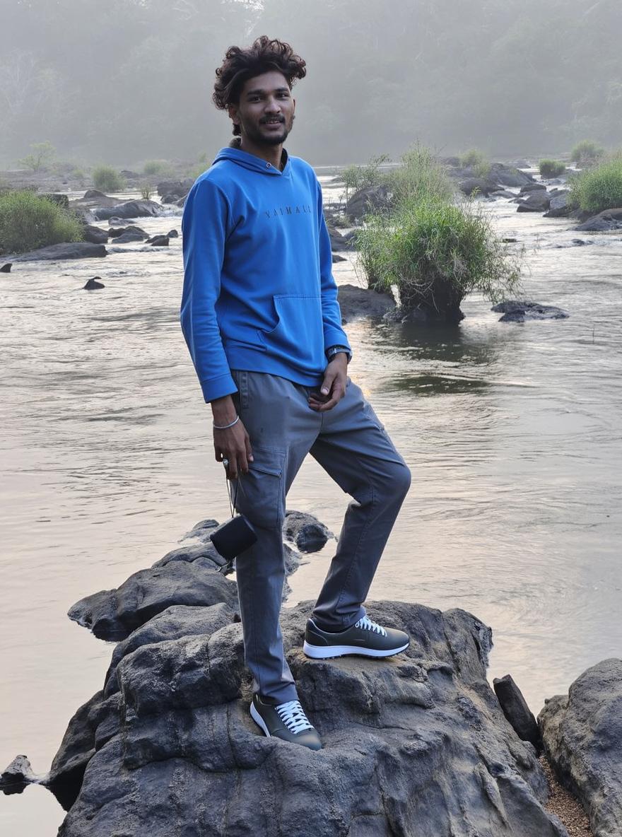 A young man standing confidently on a rocky riverbank, dressed in a light blue hoodie with stylish lettering, gray trousers, and a pair of white sneakers that contrast with his outfit