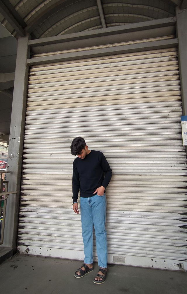 A young man standing in front of a large, closed metal shutter, wearing a black hoodie and light blue jeans