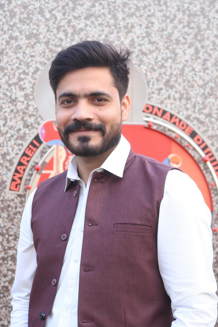 A portrait of a man wearing a traditional Indian outfit with a maroon nehru jacket over a white kurta, standing confidently against a textured wall with a colorful patterned backdrop that includes red and gold designs