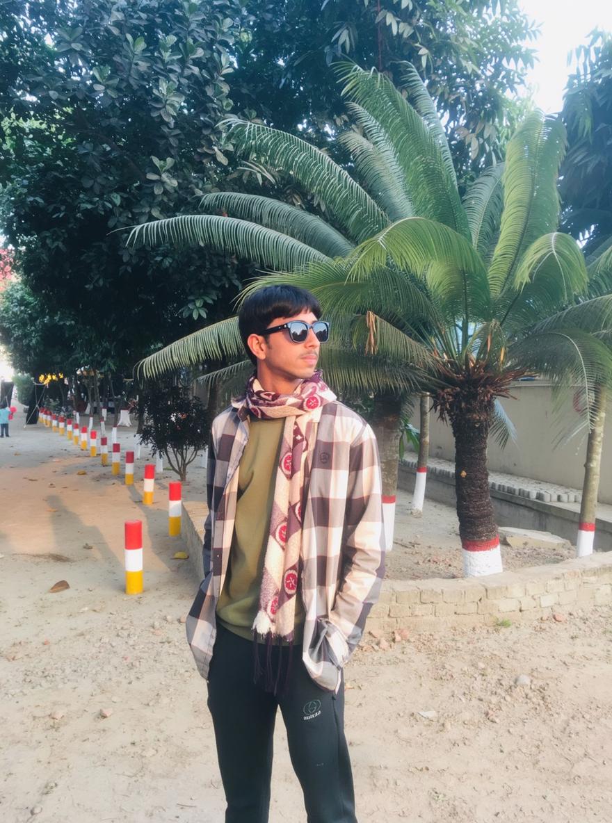 A stylish young man with long hair, standing confidently in an outdoor setting surrounded by lush green palm trees