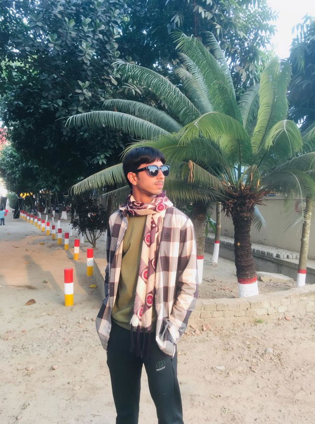 A stylish young man with long hair, standing confidently in an outdoor setting surrounded by lush green palm trees
