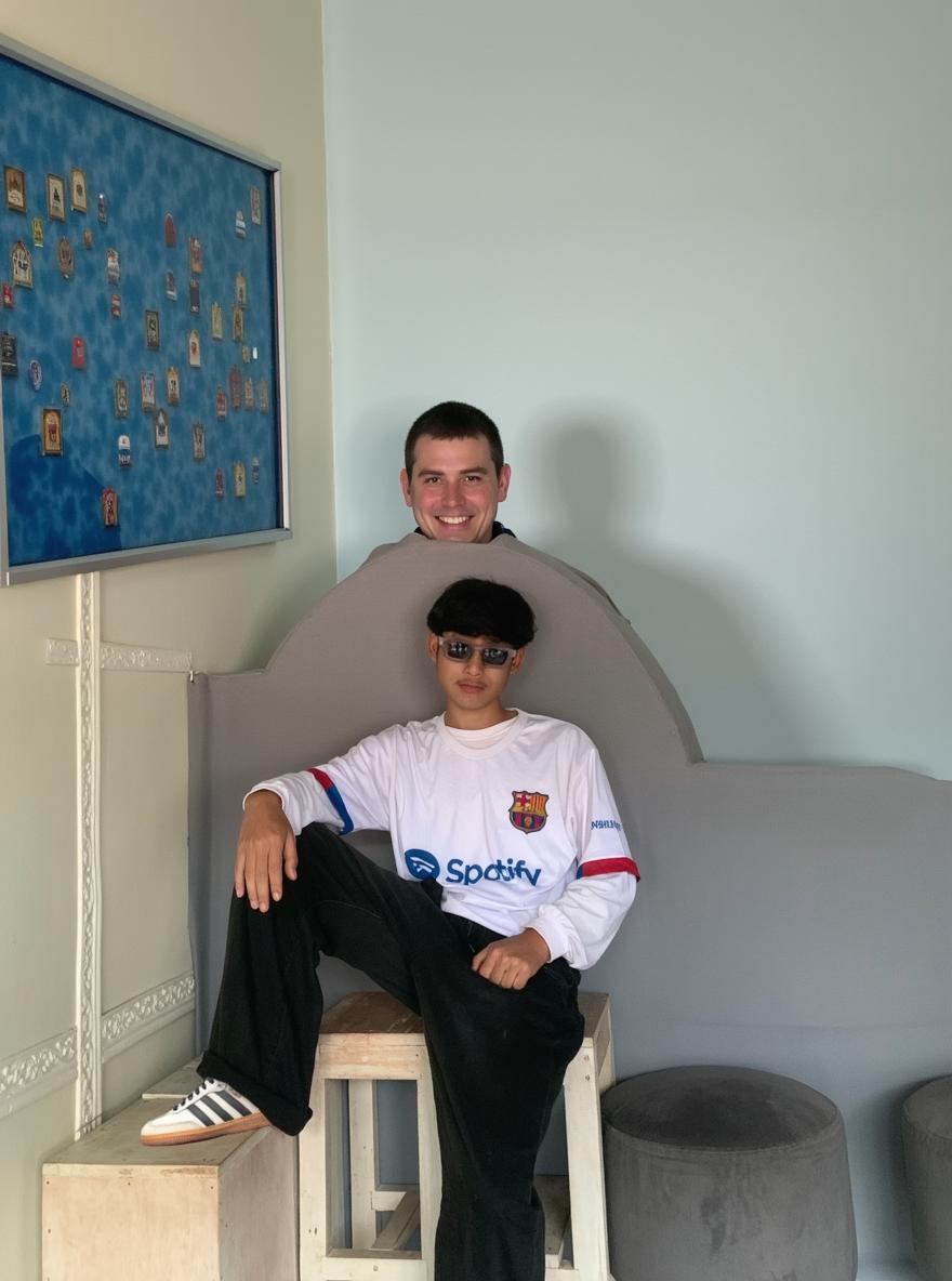 A young person sitting casually on a wooden stool, wearing a stylish, oversized white sports jersey with colorful accents, and black baggy pants