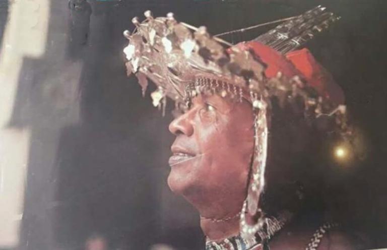 A man wearing intricate traditional tribal attire with a detailed headdress adorned with beads and shells, looking upwards in a captivating pose