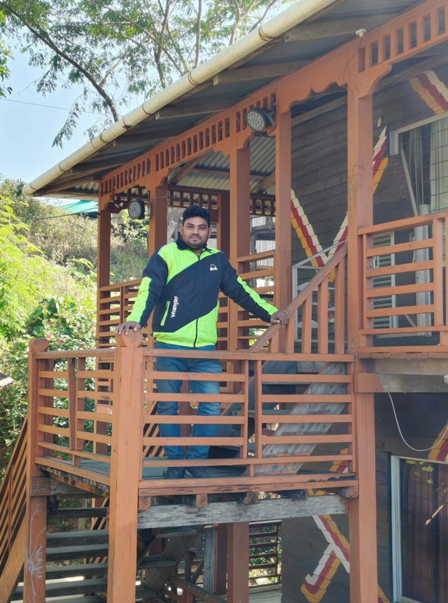 A young man standing confidently on a balcony of a wooden house, surrounded by lush greenery and trees