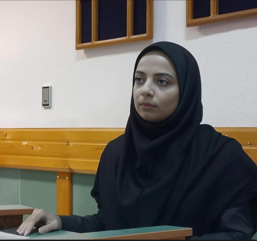 A woman in a black hijab, sitting at a desk in a modest, simple room