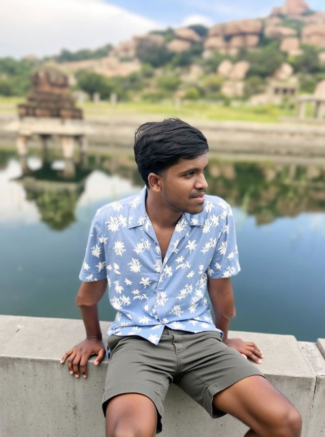 A man leaning casually against a stone wall near a serene water body, dressed in a floral blue shirt and black shorts