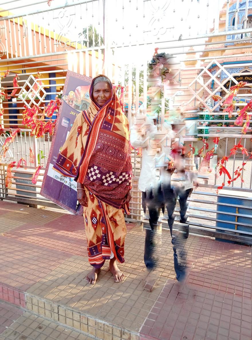 An elderly woman wrapped in a beautifully patterned shawl stands on a bustling street, surrounded by colorful decorations and vibrant banners