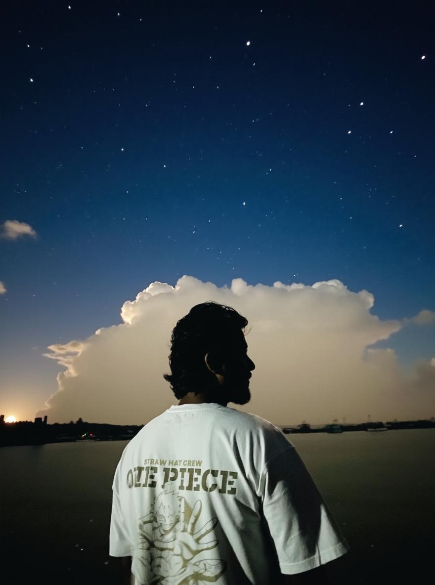 A person standing near the water at night, silhouetted against a cloudy sky illuminated by a full moon