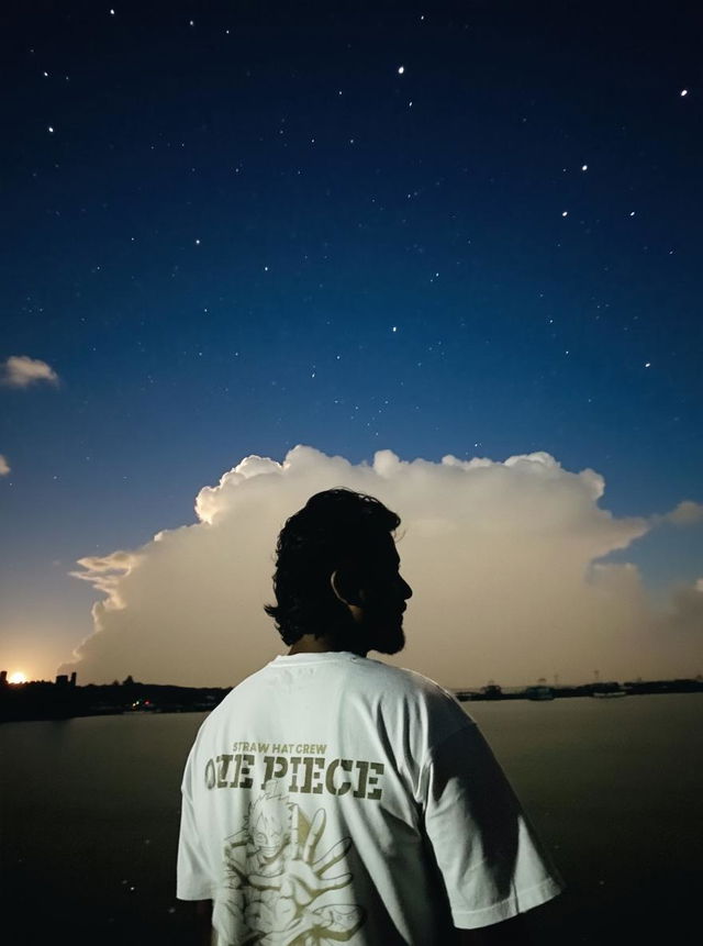 A person standing near the water at night, silhouetted against a cloudy sky illuminated by a full moon