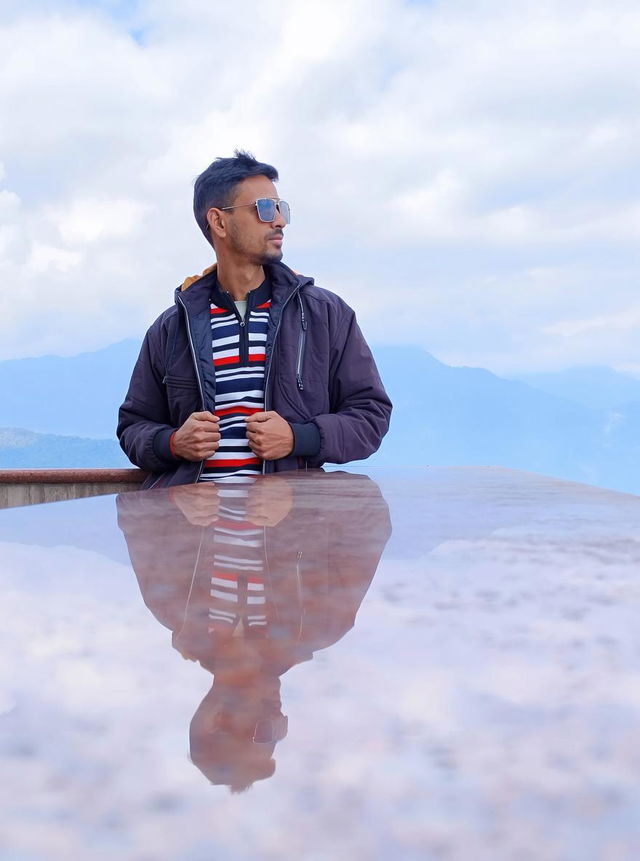 A man standing confidently at the edge of a scenic lookout, wearing a stylish jacket and sunglasses